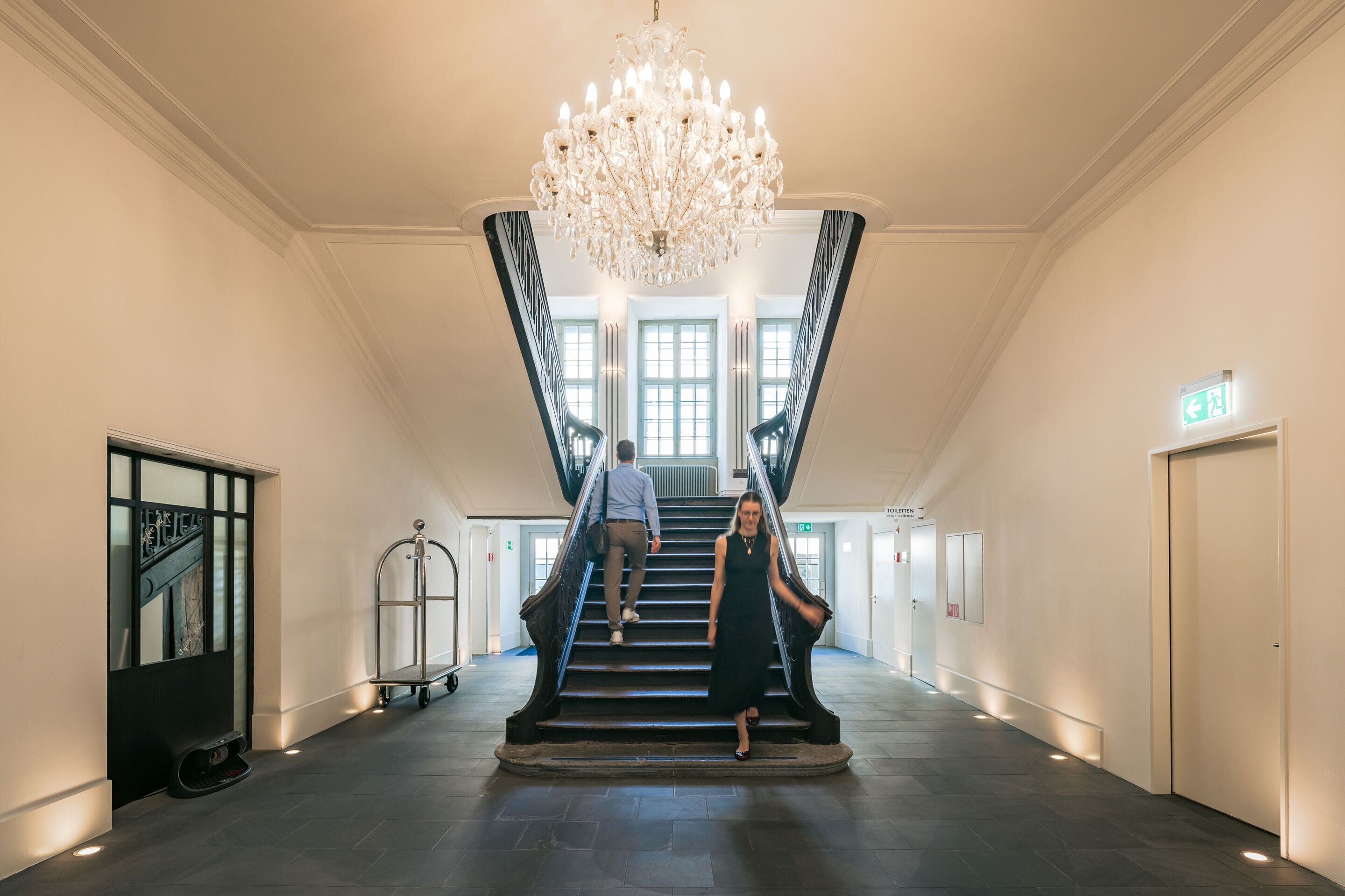 Una gran escalera con un candelabro, un hombre subiendo y una mujer bajando.