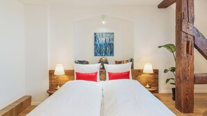 A bedroom with a large bed, two bedside tables with lamps, a wooden headboard, a painting on the wall, and a staircase to the right. - Tailormade Hotel RÜDEN Schaffhausen (Schaffhausen)