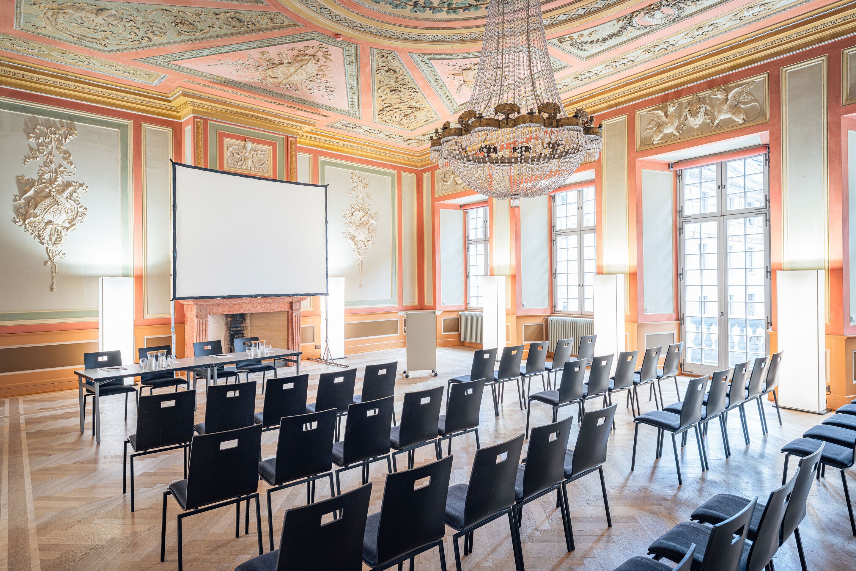 A conference room with a large screen, rows of chairs, a chandelier, and ornate ceiling decorations.