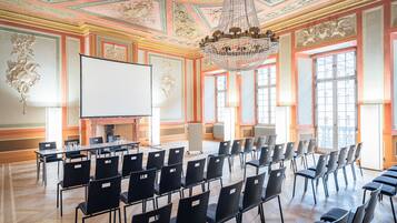 A conference room with a large screen, rows of chairs, a chandelier, and ornate ceiling decorations.