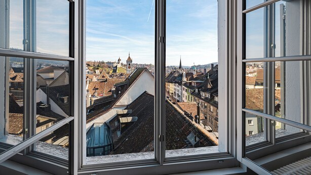 A view through a window with a clear sky, featuring a cityscape with multiple buildings and a tower. - Tailormade Hotel RÜDEN Schaffhausen (Schaffhausen)