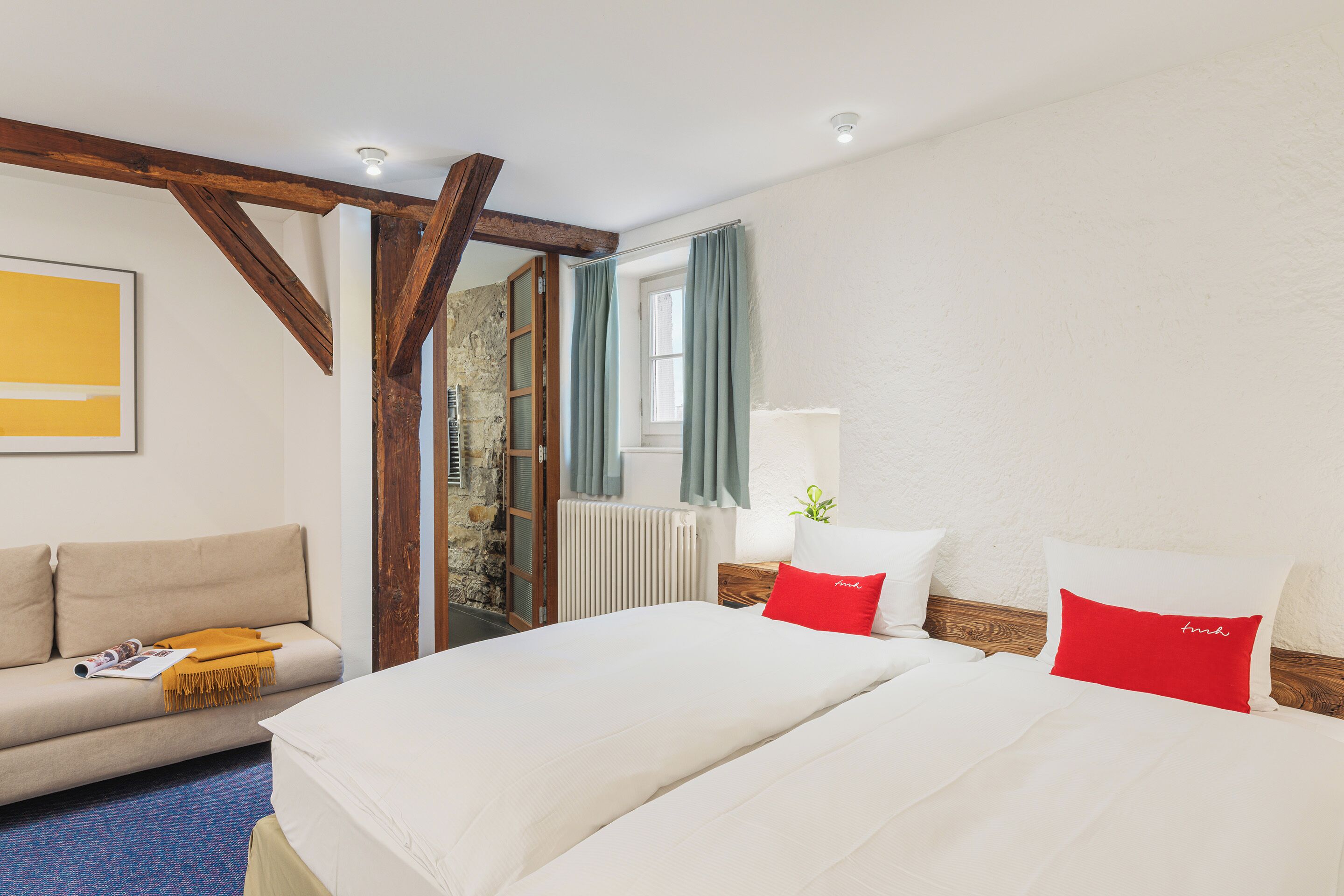Habitación de hotel con cama, sofá, ventana con cortinas, radiador y viga de techo de madera.