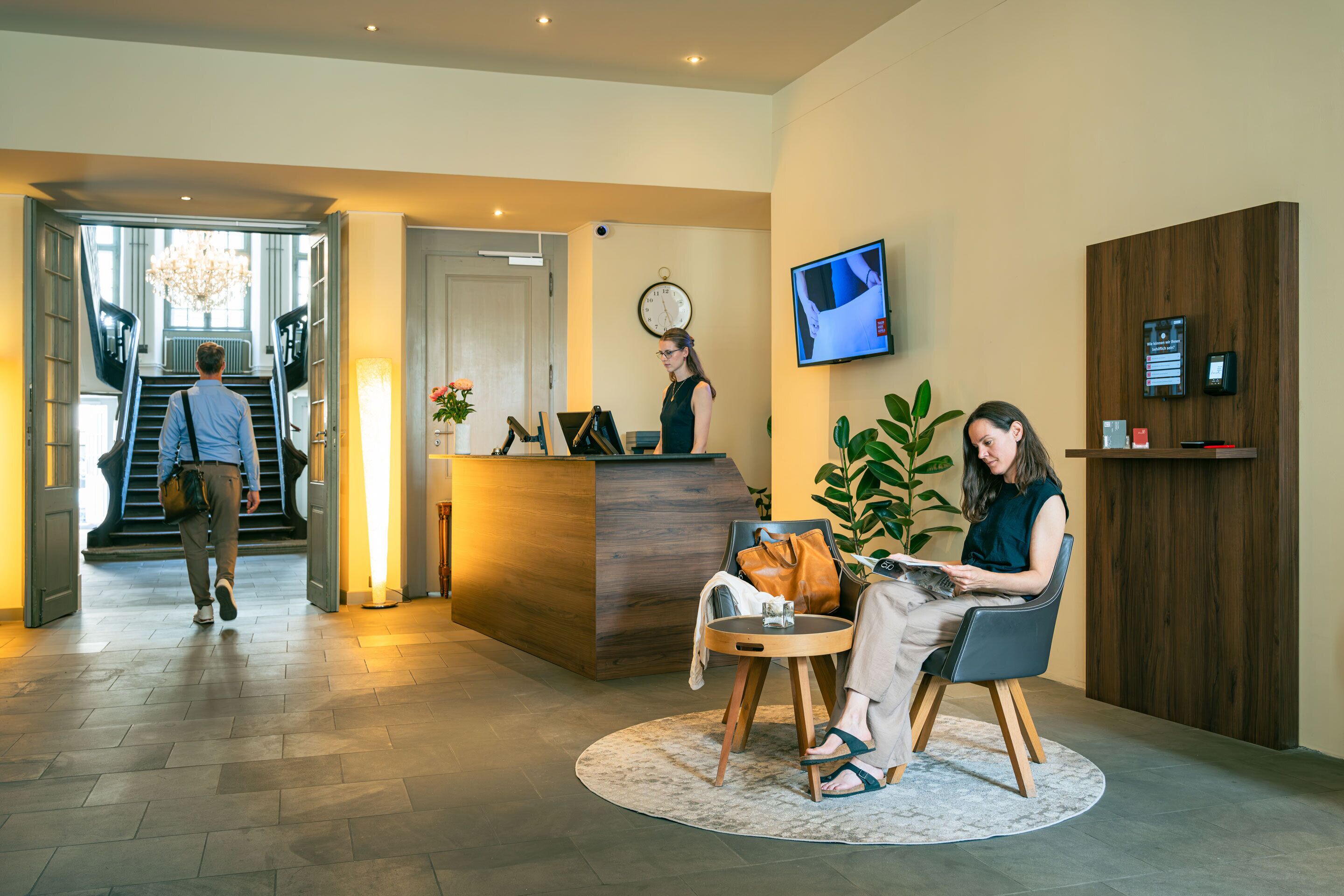 A hotel reception area with a reception desk, a clock, a TV screen, and a staircase leading to an upper floor.