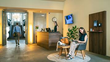 A hotel reception area with a reception desk, a clock, a TV screen, and a staircase leading to an upper floor.