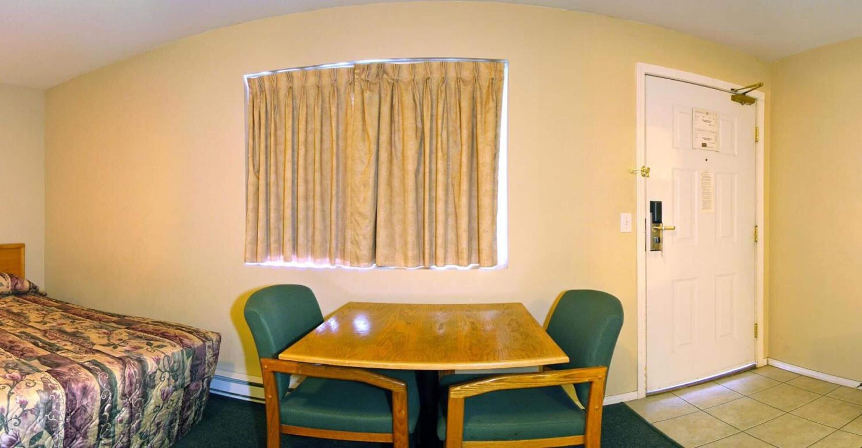 A hotel room with a bed, a table with two chairs, a window with curtains, and a door with a lock.