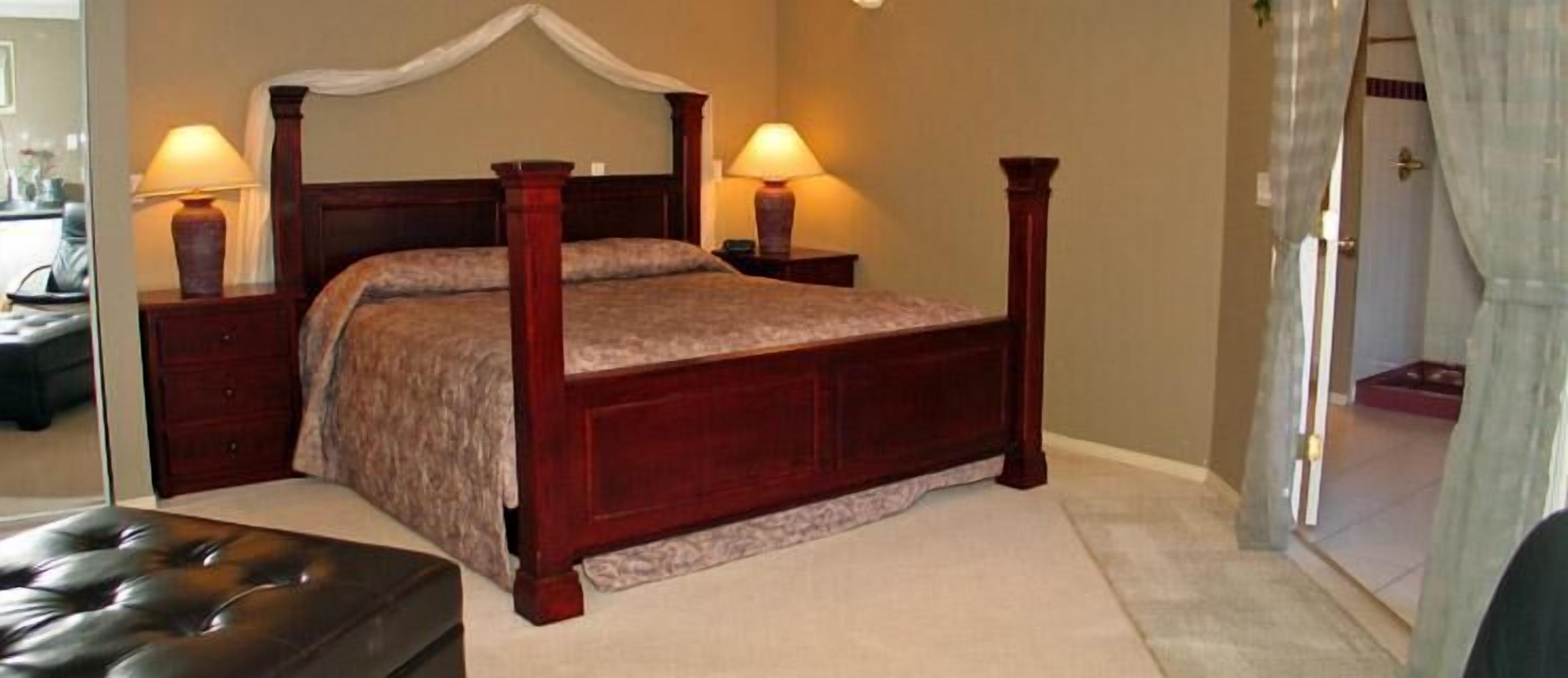 A four-poster bed with a canopy, two bedside lamps, a wooden nightstand, and a tufted leather ottoman.