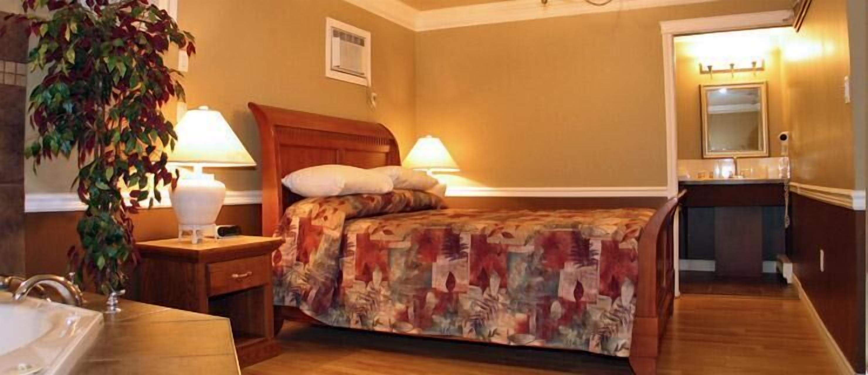 Hotelkamer met een bed, bedlampen, nachtkastje, een plant, een badkamer met spiegel en een houten vloer.