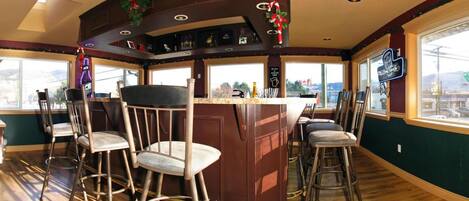 A bar area with a wooden bar counter, bar stools, and a view of the outside through large windows.