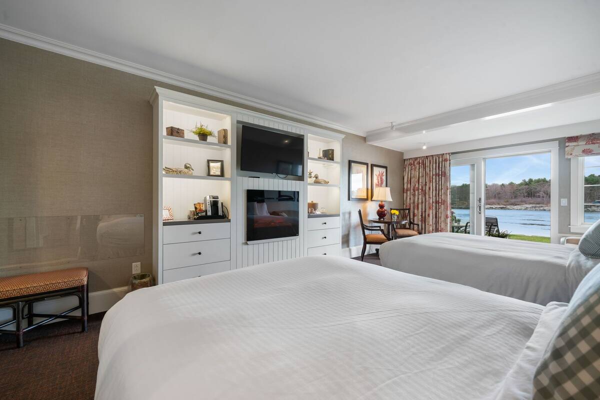 A hotel room with two beds, a TV, a desk with a chair, and a view of a lake.