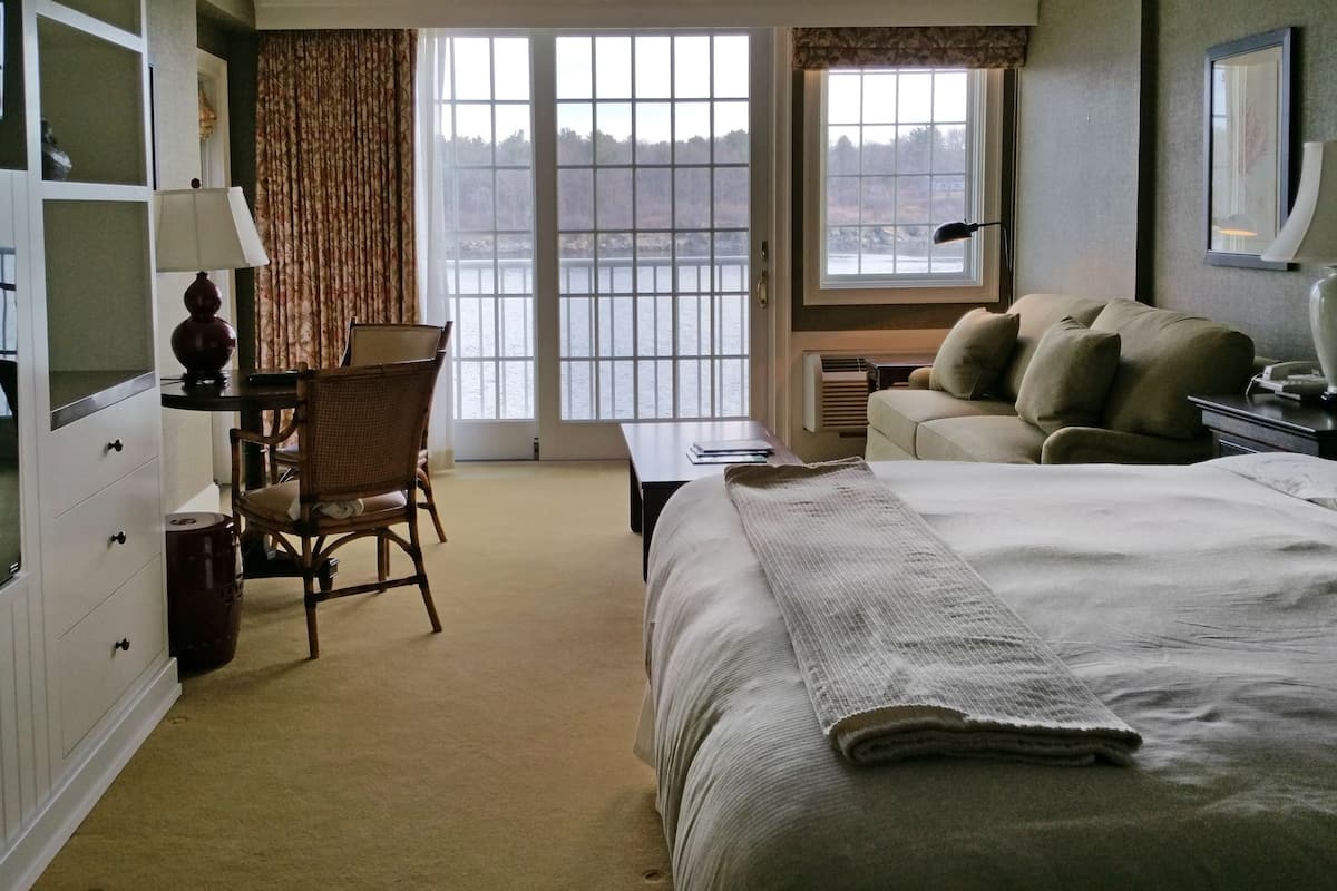 A hotel room with a large bed, a chair, a desk, and a view of a lake through the windows.