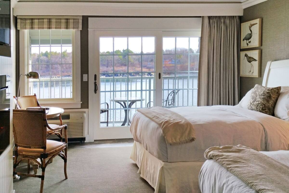 A hotel room with a bed, a desk with a lamp, a chair, and a view of a body of water through a glass door.