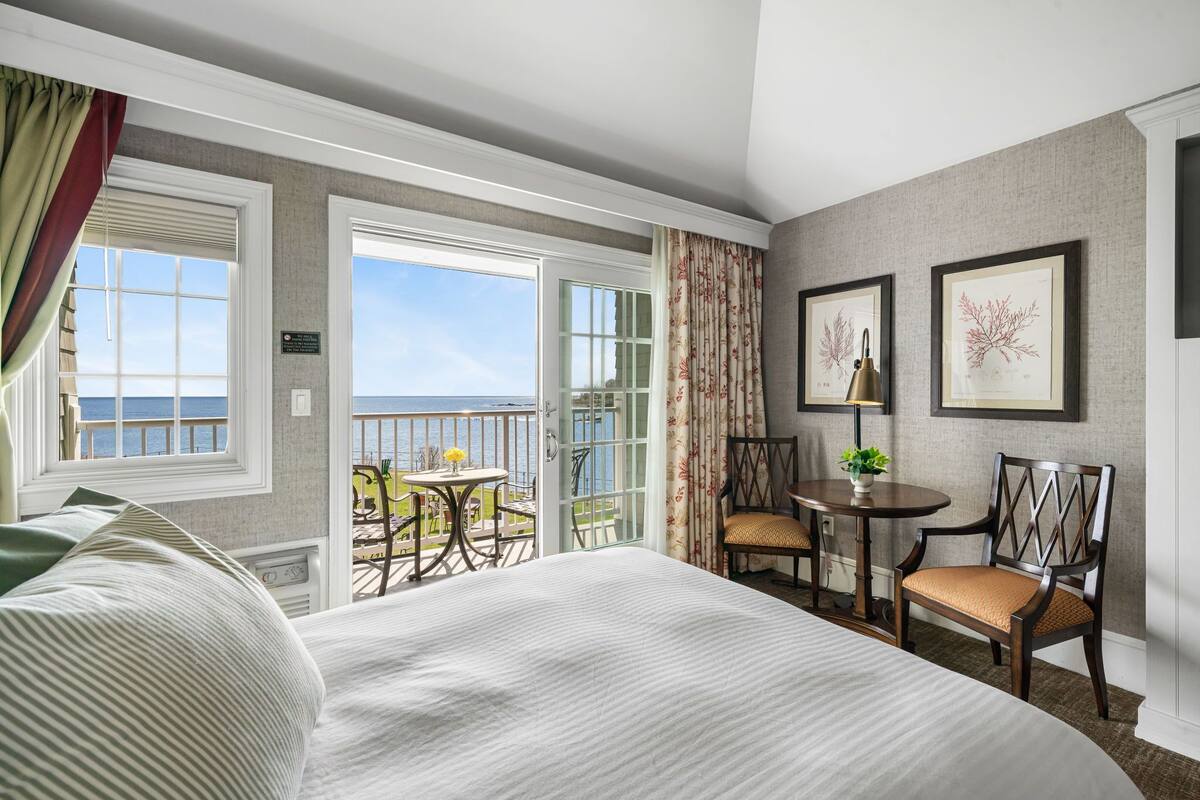 A hotel room with a large bed, a balcony with a view of the sea, a small table with chairs, and framed botanical prints on the wall.