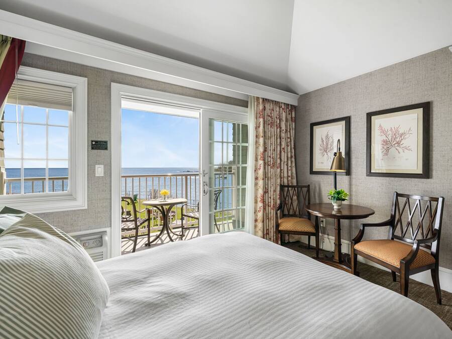 Habitación de hotel con una cama grande, un balcón con vistas al mar, una pequeña mesa con sillas e impresiones botánicas enmarcadas en la pared.