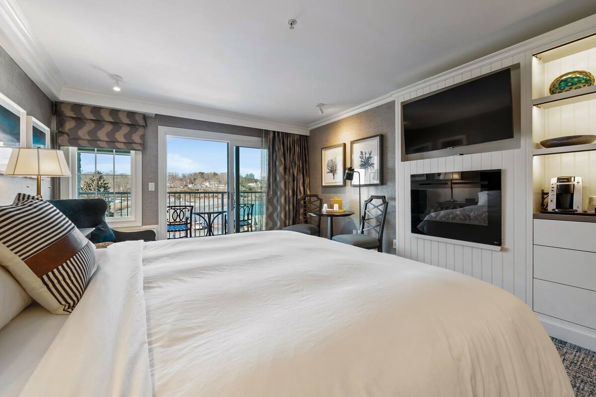 A hotel room with a large bed, a TV, a balcony with outdoor seating, and a kitchenette.