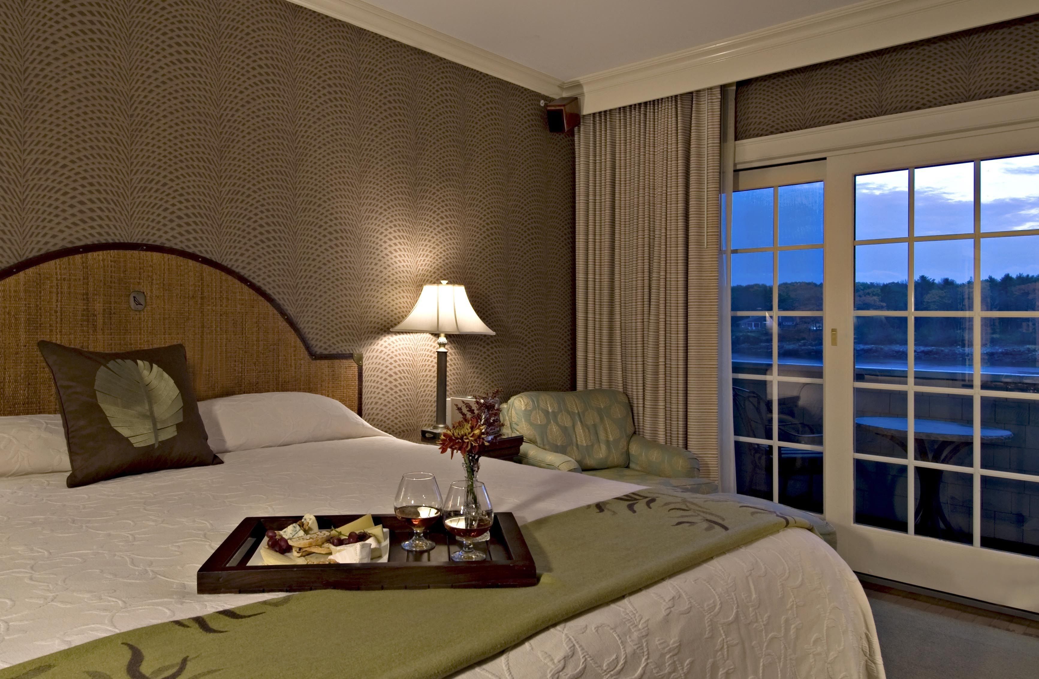 Une chambre d’hôtel avec un grand lit, une table de chevet avec une lampe, un plateau avec de la nourriture et des boissons, et une vue sur l’extérieur par une fenêtre.