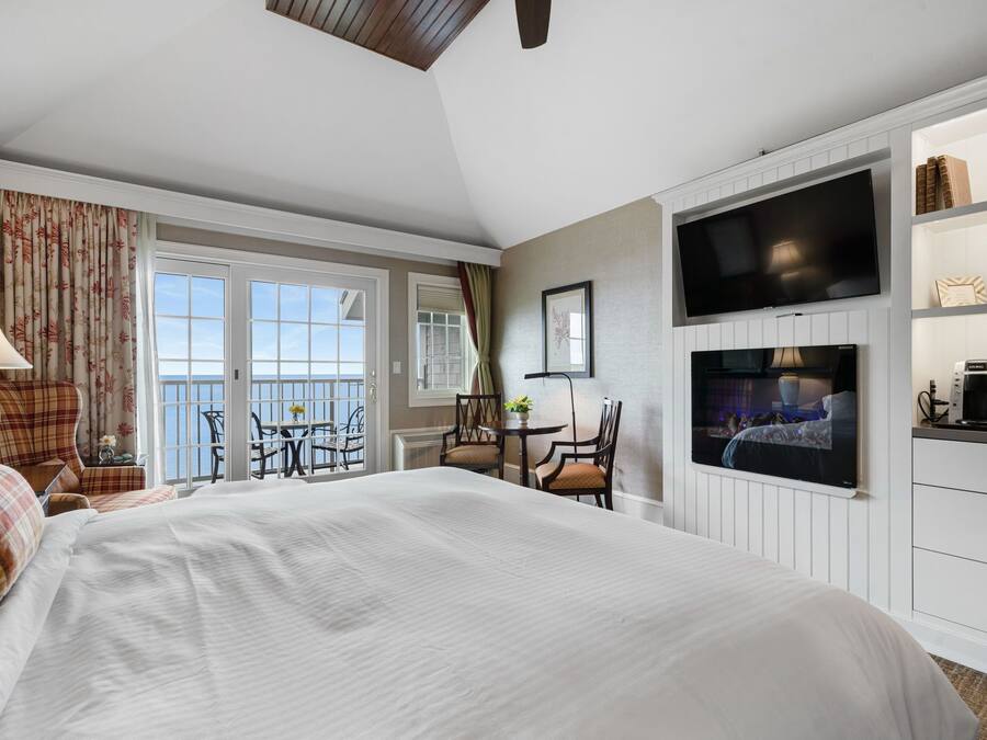 A hotel room with a large bed, a TV mounted on the wall, a balcony with chairs, and a dining area.