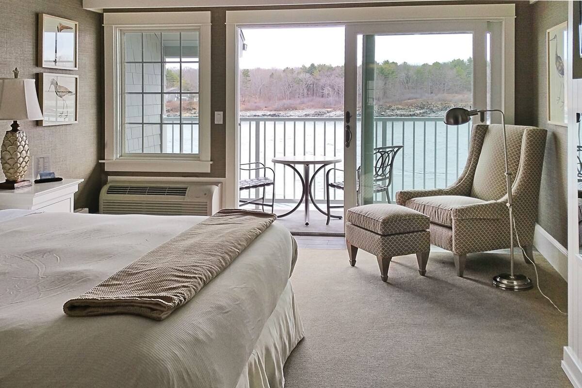 A hotel room with a large bed, a chair, a table, and a balcony with a view of the water.