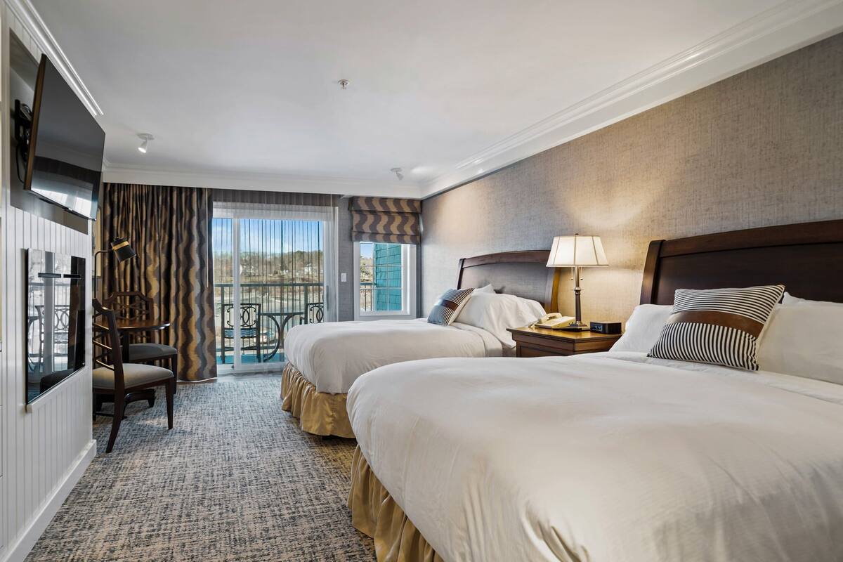A hotel room with two beds, a TV, a desk with a lamp, and a view of the outdoors.