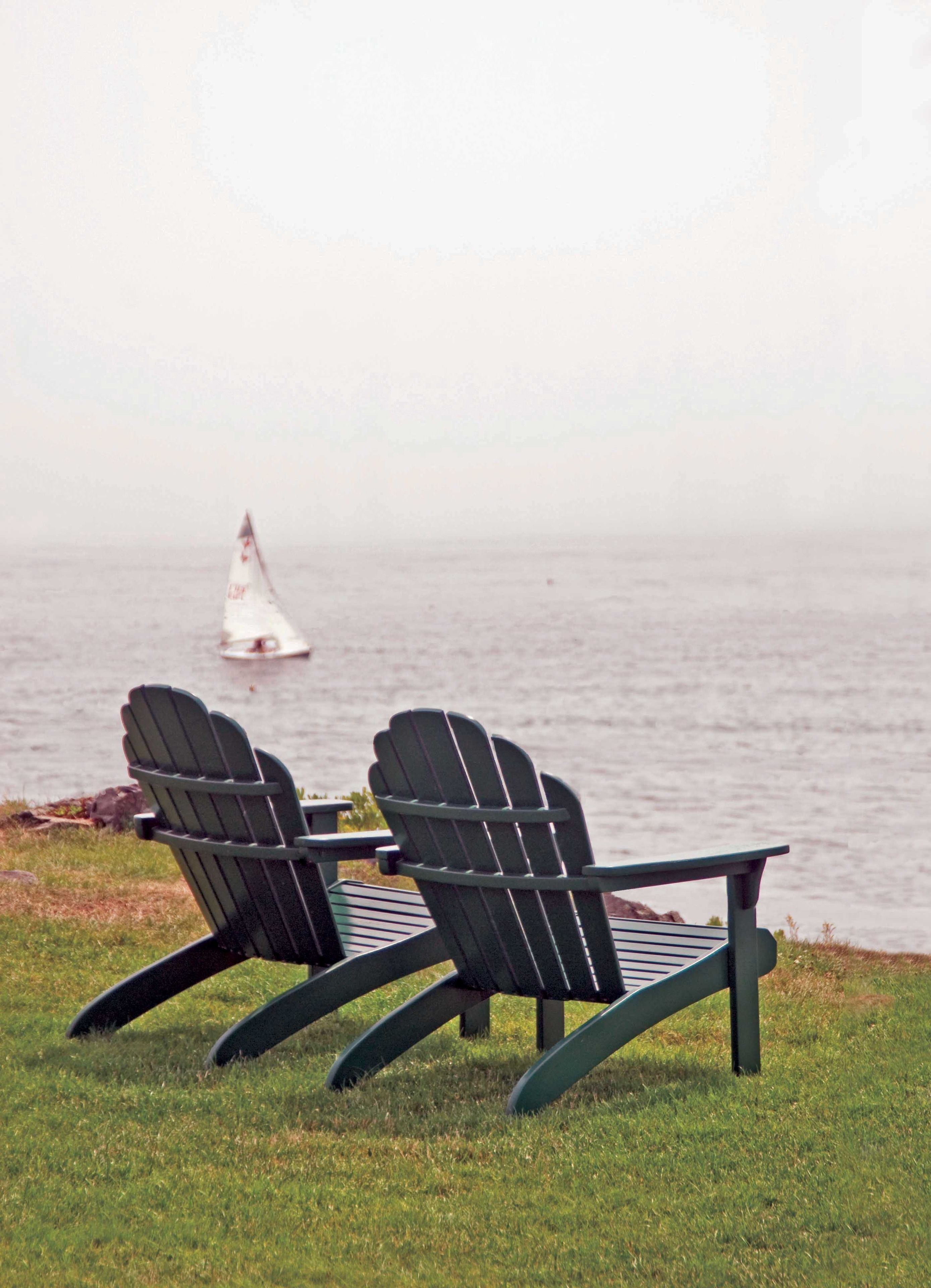 Two Adirondack chairs facing a body of water with a sailboat in the distance.
