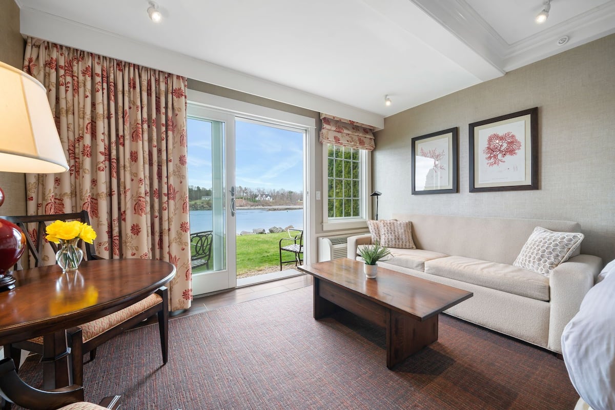 A living room with a sofa, coffee table, and a dining table with chairs. There are two framed pictures on the wall and a window with curtains.