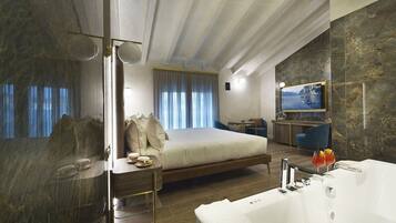 Ein modernes Hotelzimmer mit einem großen Bett, einer separaten Sitzecke mit Fernseher, einem Badezimmer mit Badewanne und Blick auf das Meer durch das Fenster.