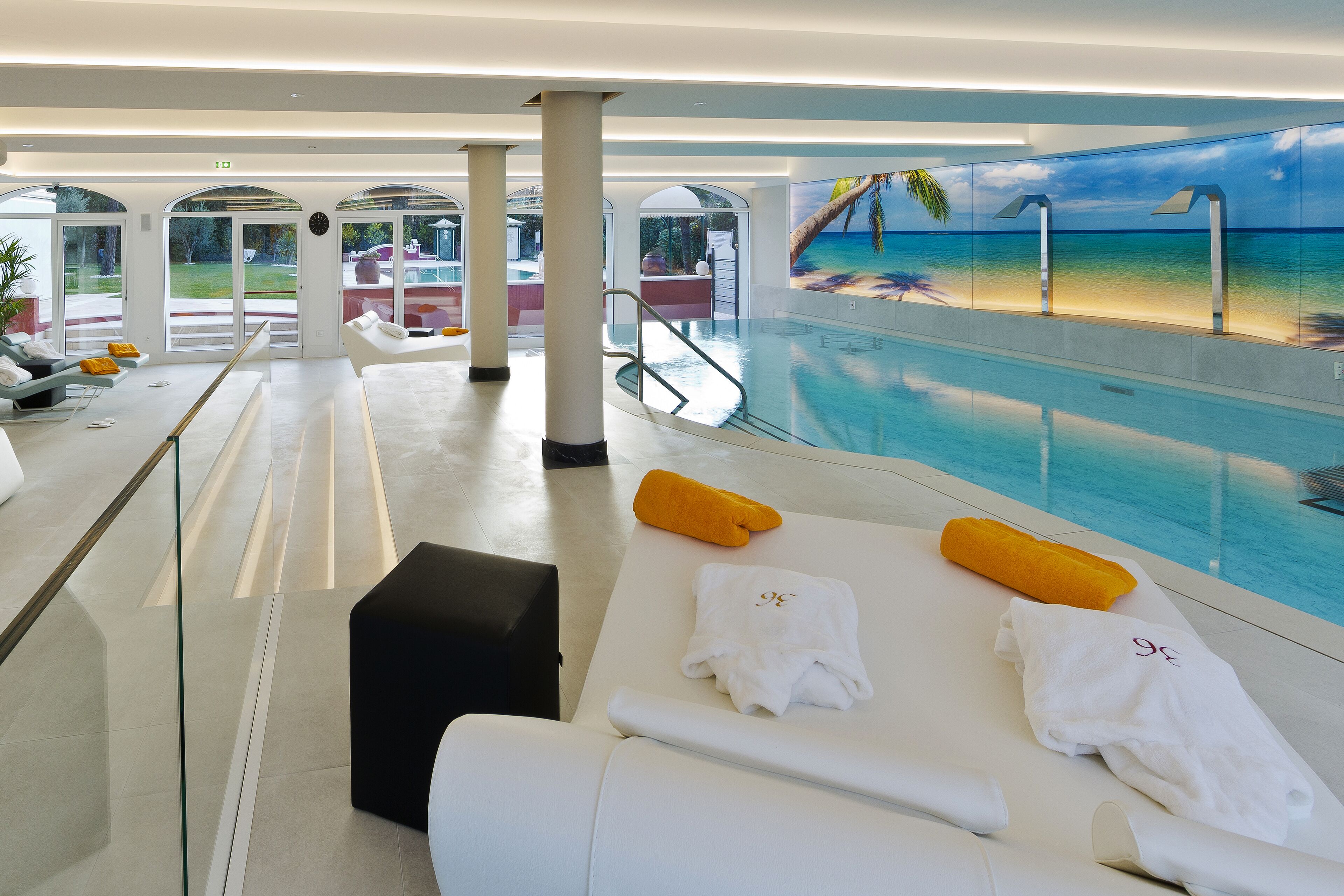 Un área de piscina interior moderna con sillones de descanso, barandilla de vidrio y un mural de una playa.