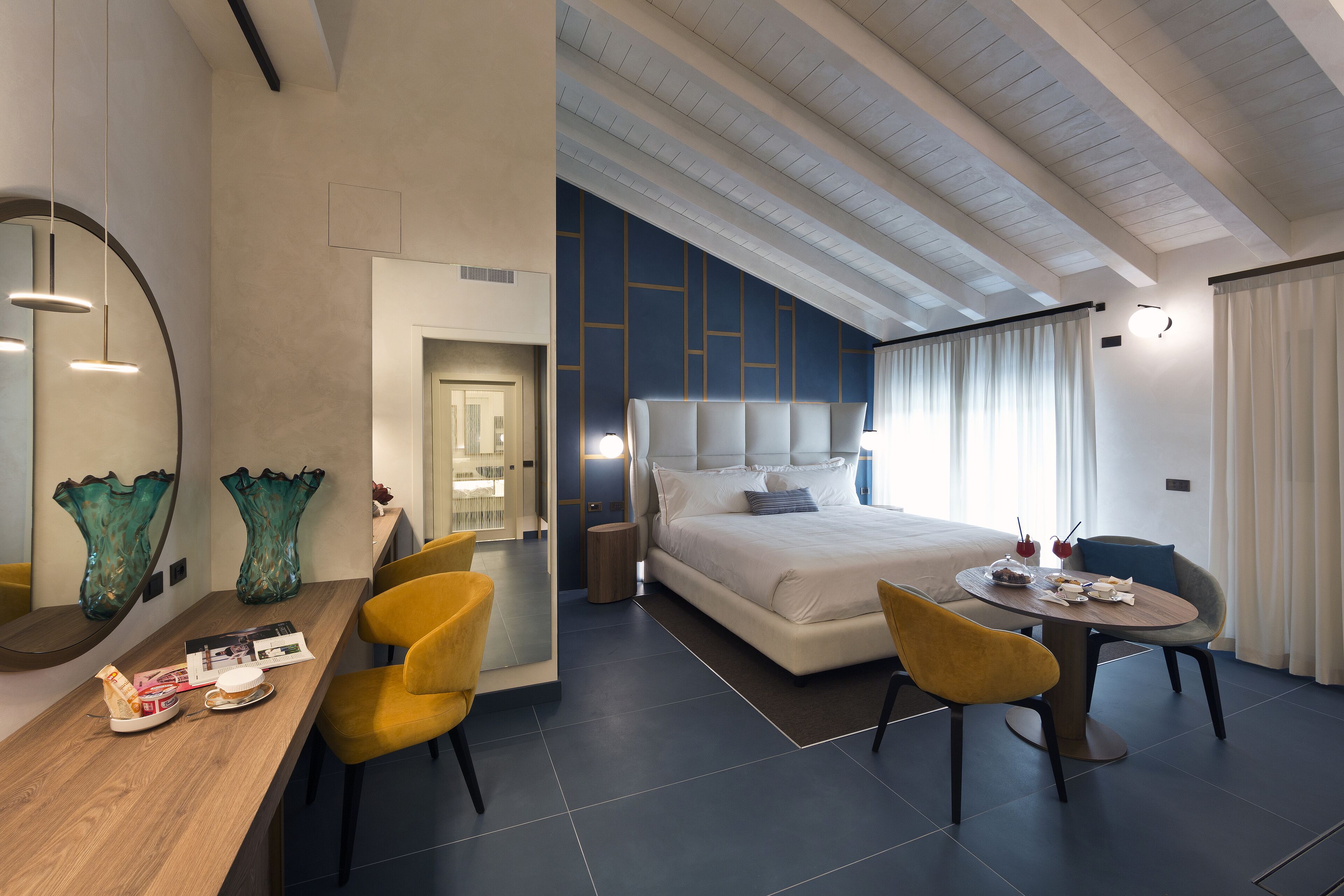 A modern hotel room with a large bed, a dining table, yellow chairs, a wooden desk, and decorative vases.