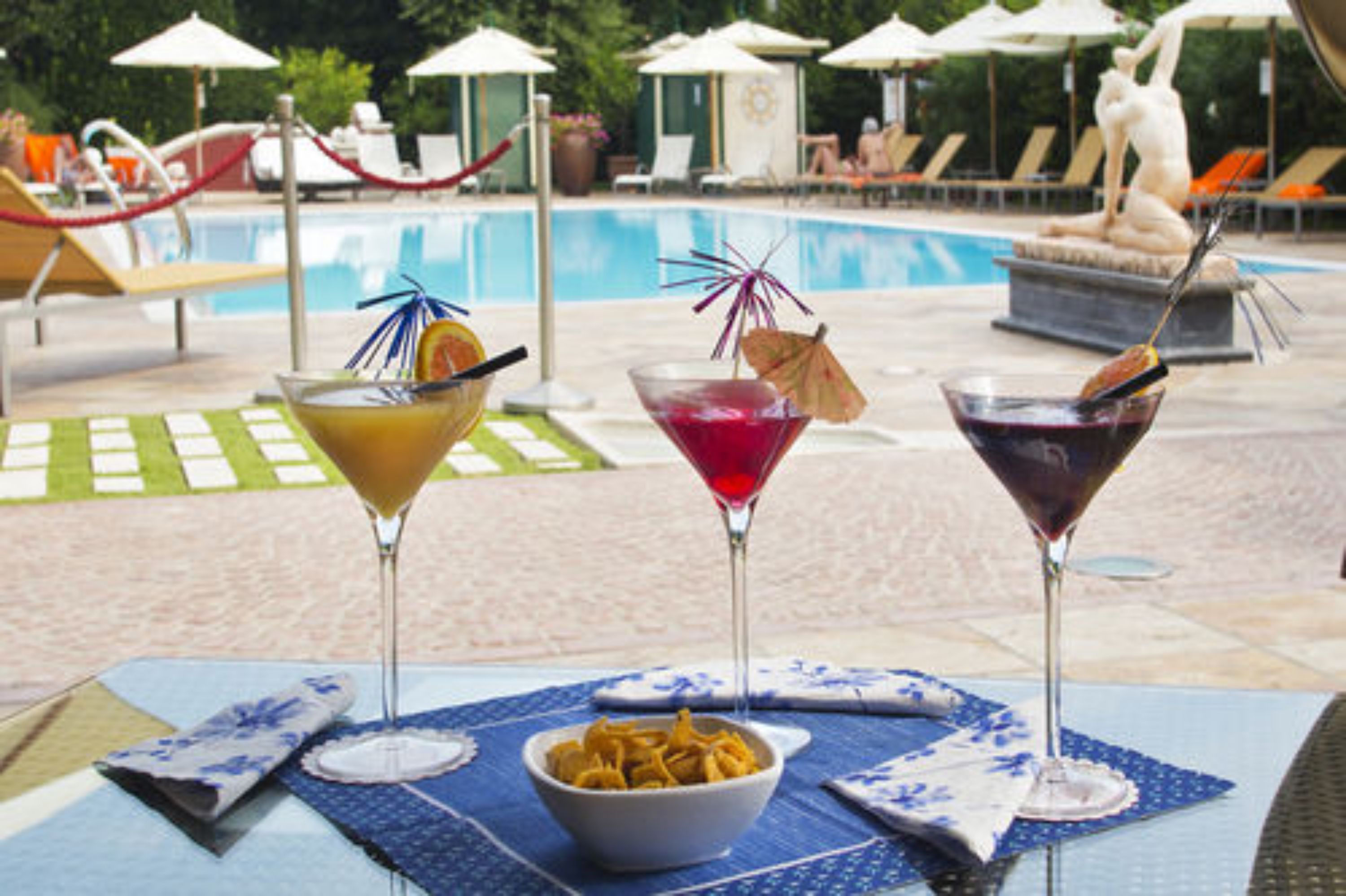 A poolside table with three martini glasses, a bowl of snacks, and a statue in the background.