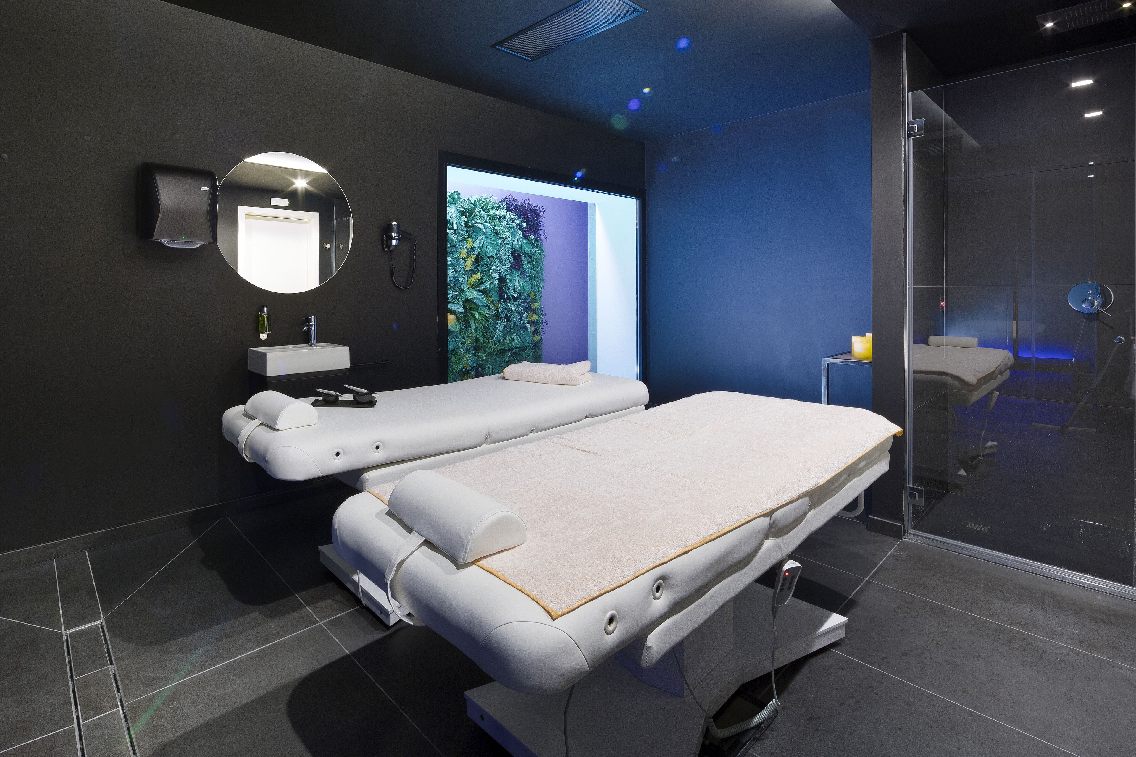 Un moderno spa con dos camillas de masaje, un espejo montado en la pared y una zona de ducha con paredes de vidrio.