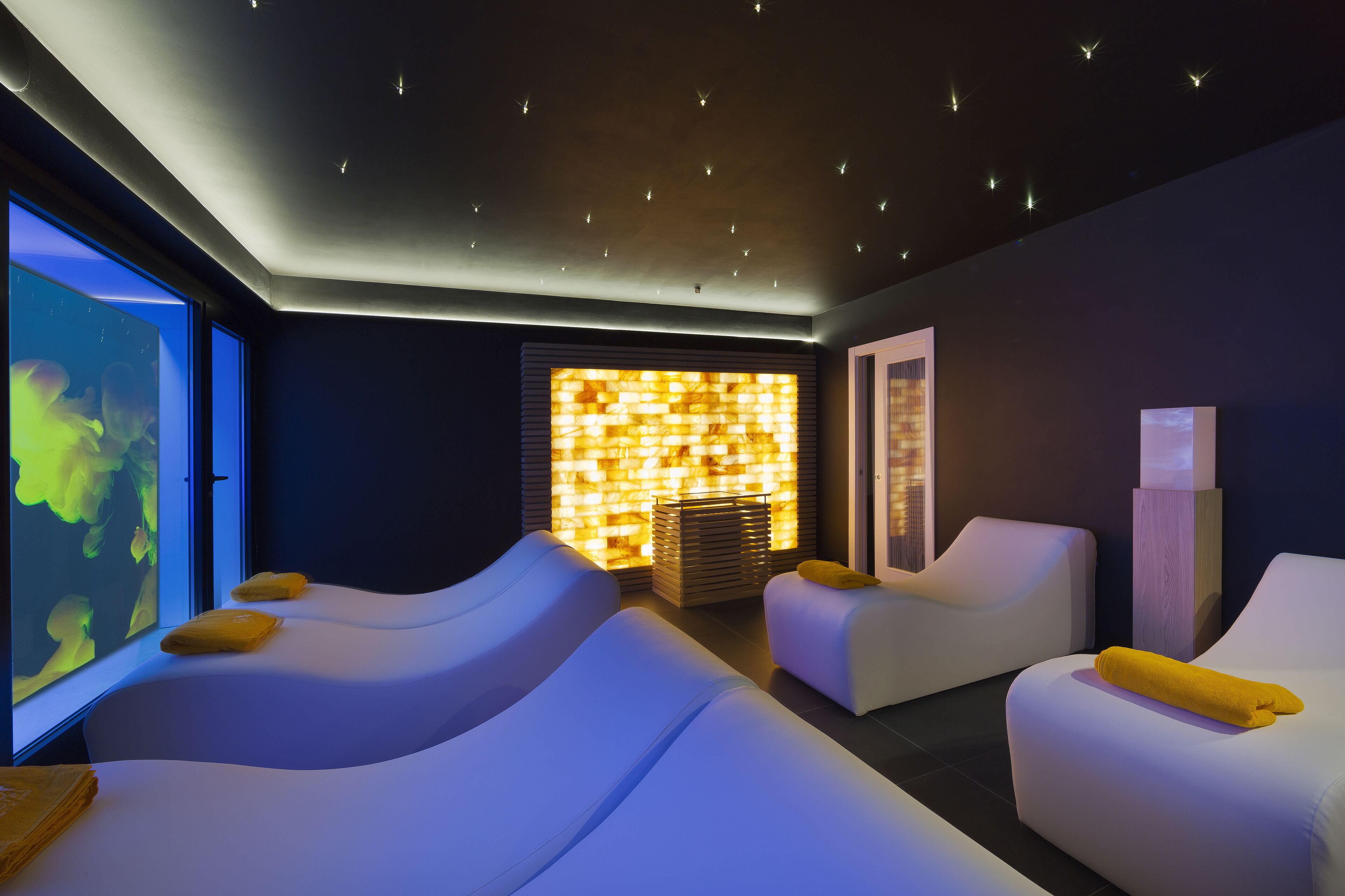 A spa room with two massage tables, a wall with a textured light panel, and a large window with a view.