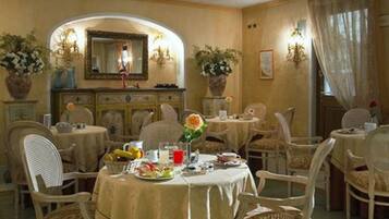 Un comedor con varias mesas redondas cubiertas con manteles color beige, rodeado de sillas de mimbre.
