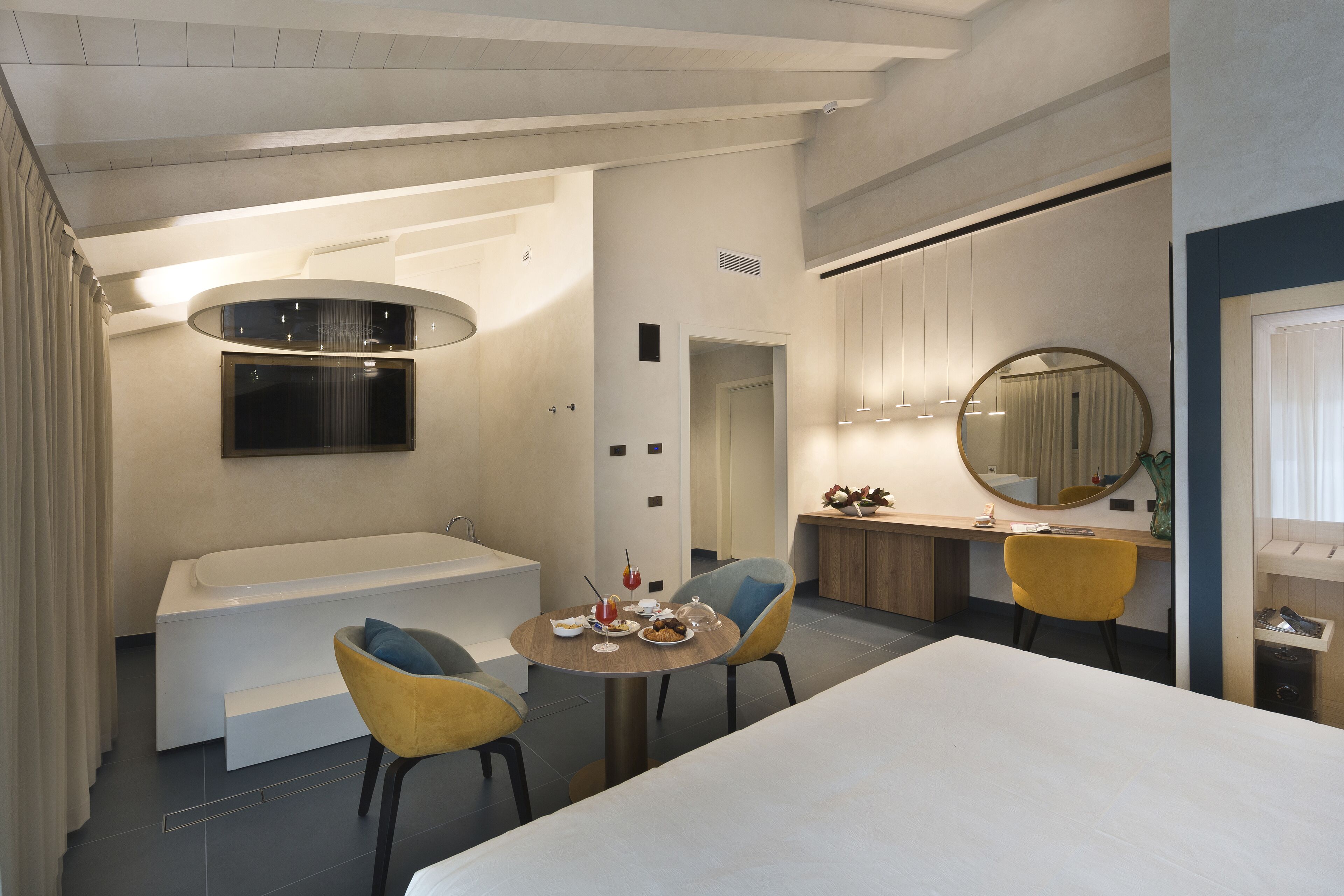 A modern hotel room with a large bed, a round dining table, two chairs, a bathtub, a mirror, and a television.