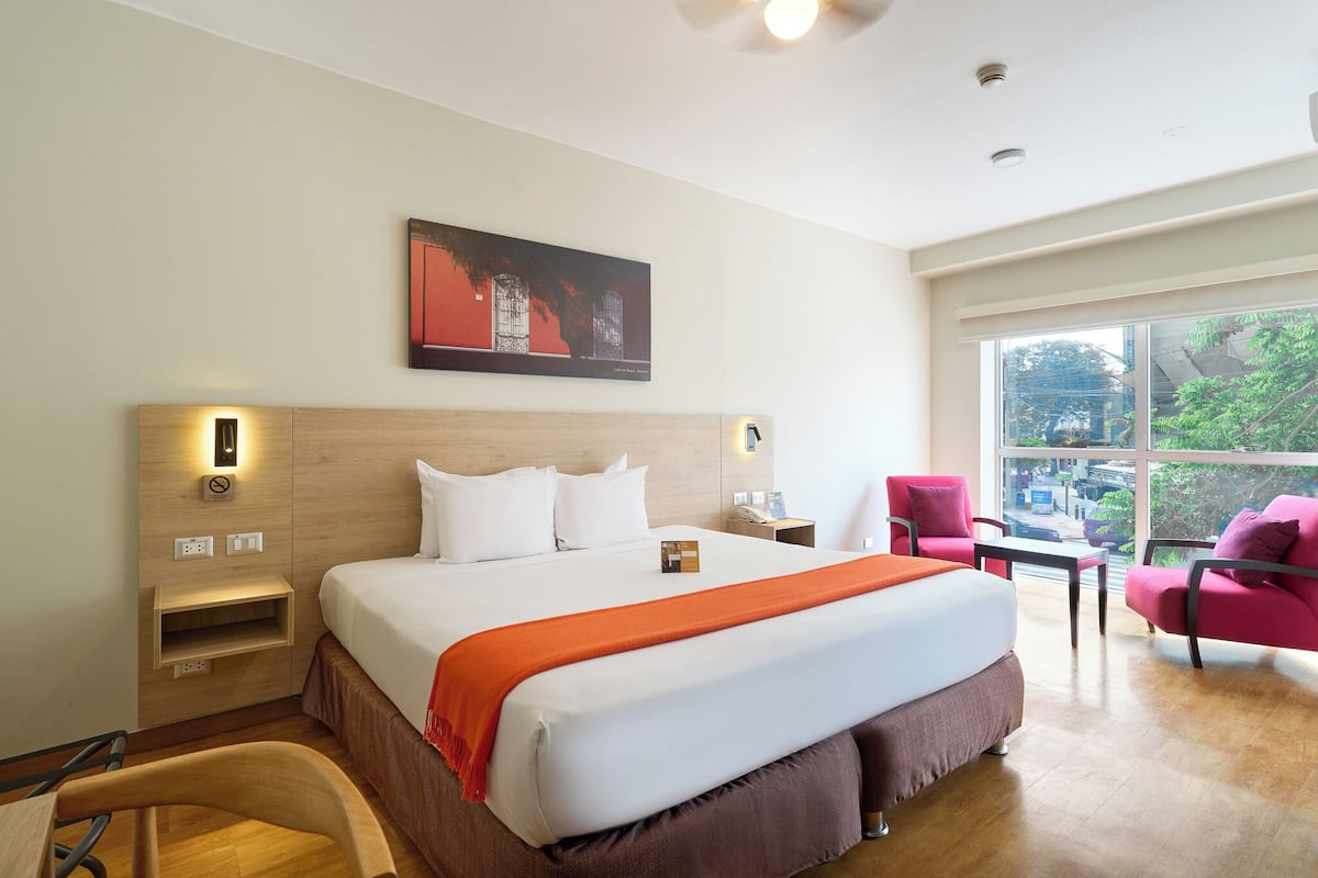 A hotel room with a large bed, a desk, a chair, a window with a view, and a painting on the wall.