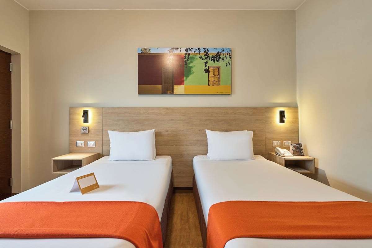 A hotel room with two beds, a wooden headboard, bedside tables with lamps, a painting on the wall, and a small card on the left bed.