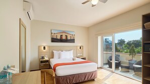 Habitación de hotel con cama, escritorio, silla, dispensador de agua, ventilador de techo y una ventana grande con vistas a edificios y árboles.