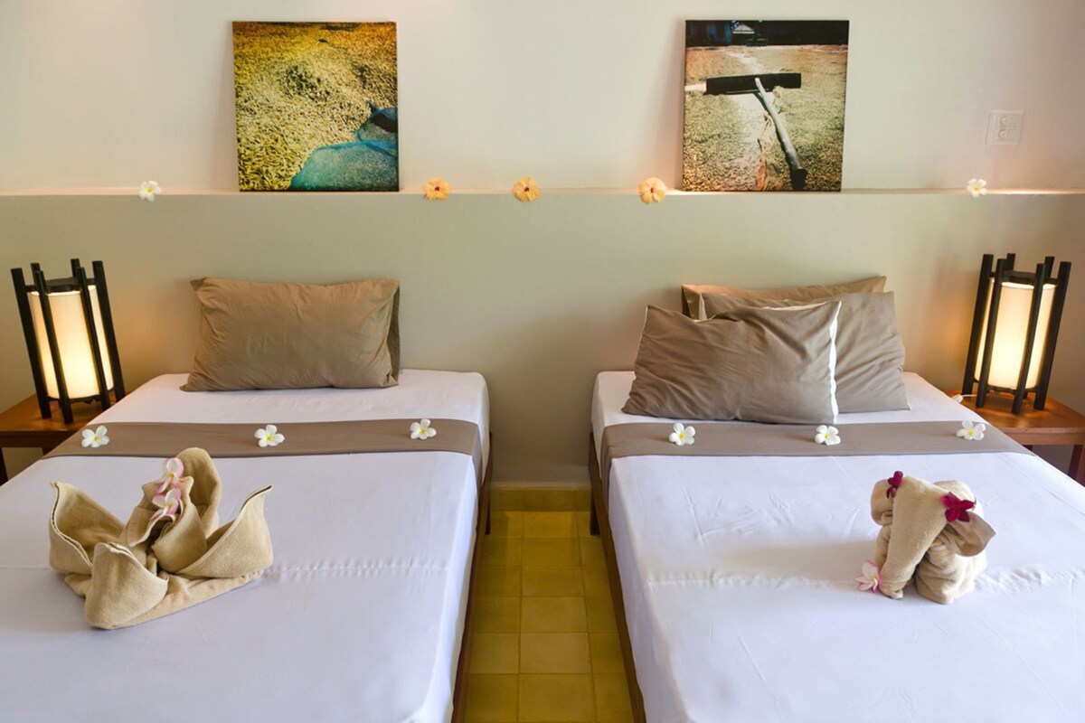 Dos camas individuales con ropa de cama color beige, almohadas y toallas decorativas con flores.