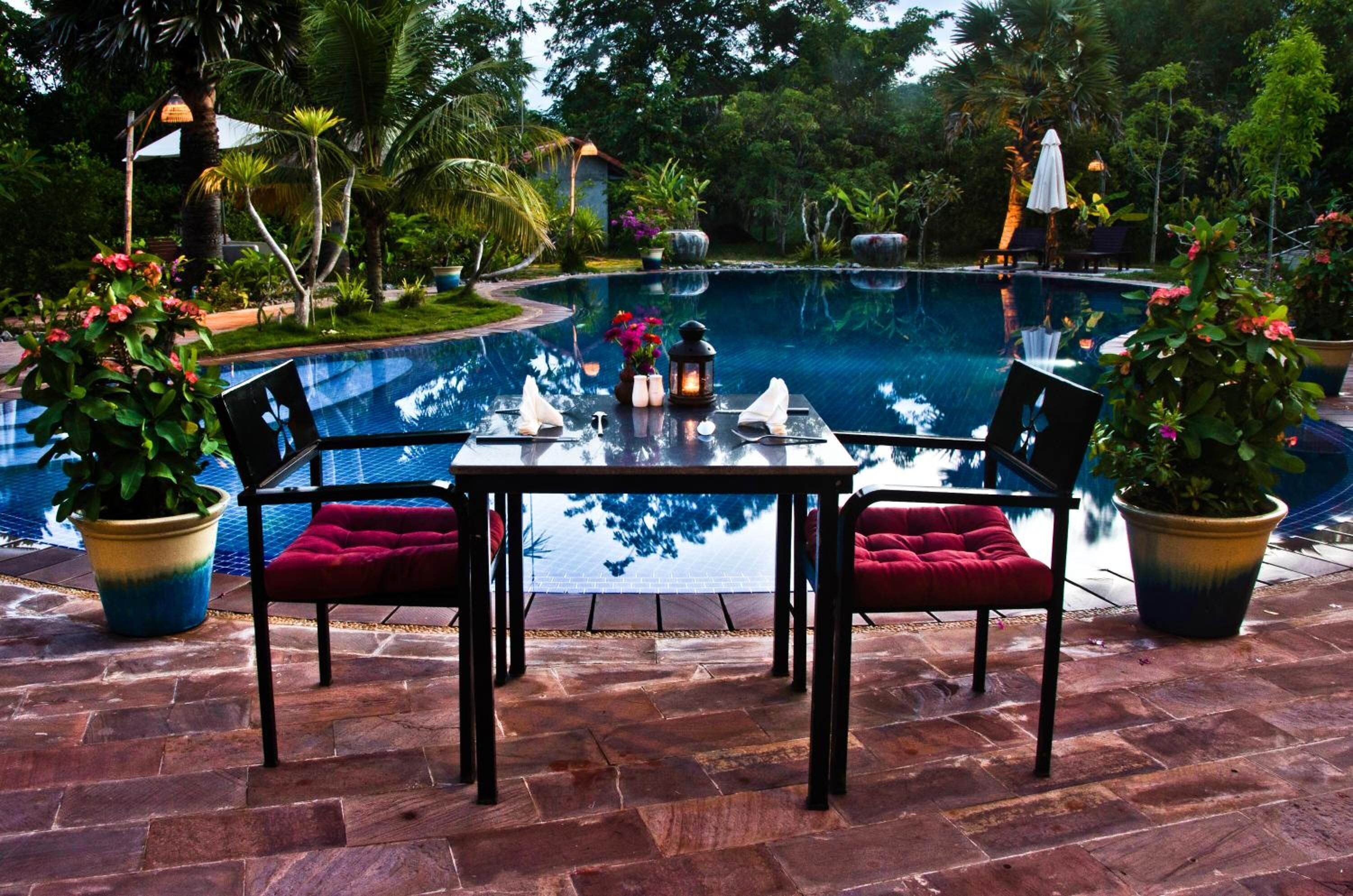 Un área de comedor junto a la piscina, con mesa y sillas, rodeada de exuberante vegetación y una piscina cristalina.