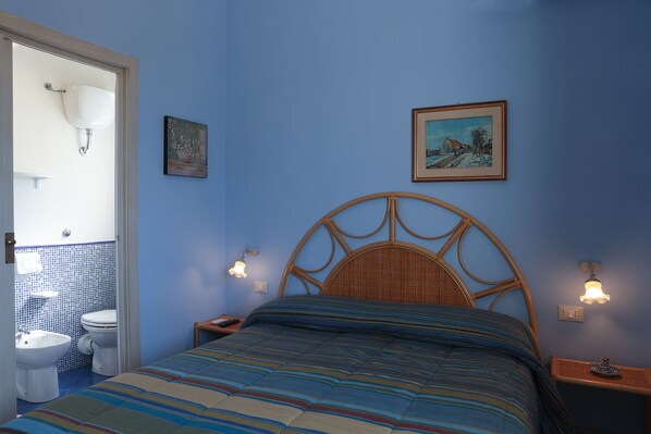 A bedroom with a bed, a nightstand, a wall-mounted lamp, a framed picture, and a view of the bathroom. - Villa dei Giardini (Agrigento)