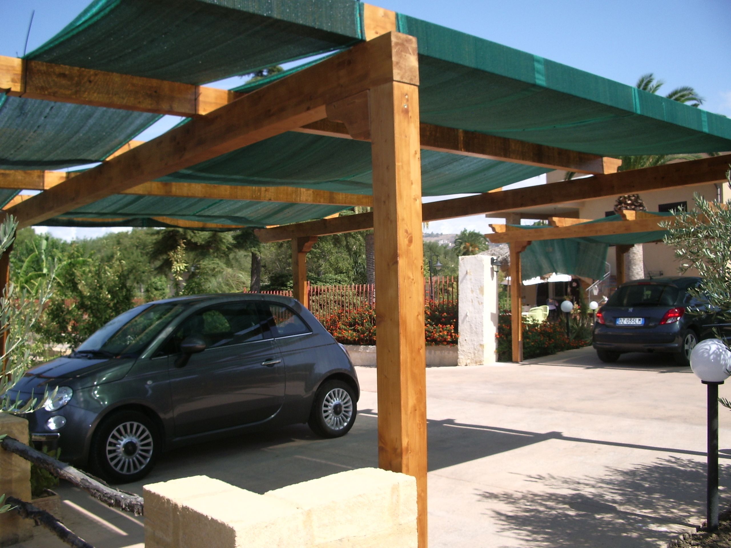 Un coche aparcado bajo una pérgola de madera con techo verde, rodeado de una zona pavimentada y una valla con flores.