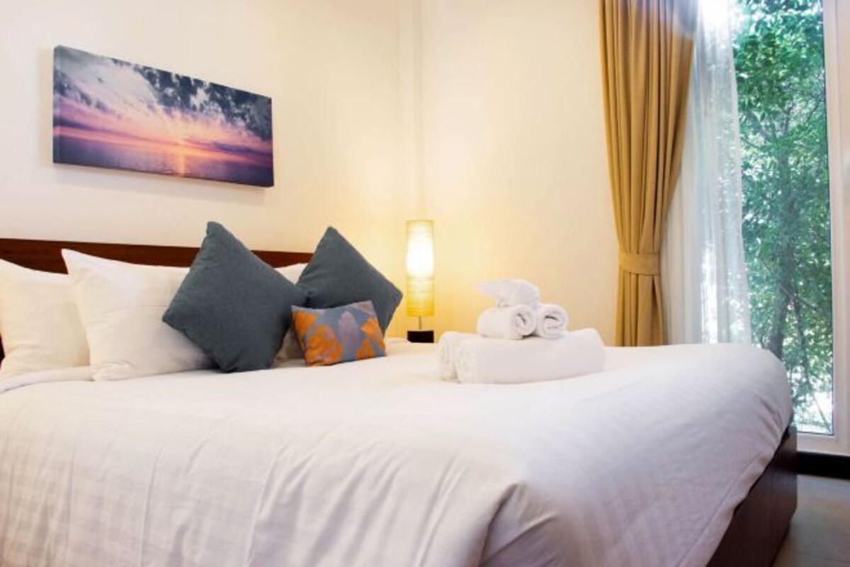 Une chambre d’hôtel avec un lit, des oreillers, une lampe et une vue sur les arbres par la fenêtre.