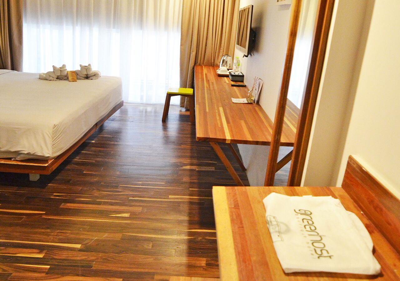 A hotel room with a wooden floor, a bed with a white bedspread, a wooden desk with a chair, a small table with a towel, and a television on the wall.