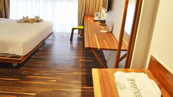 A hotel room with a wooden floor, a bed with a white bedspread, a wooden desk with a chair, a small table with a towel, and a television on the wall.