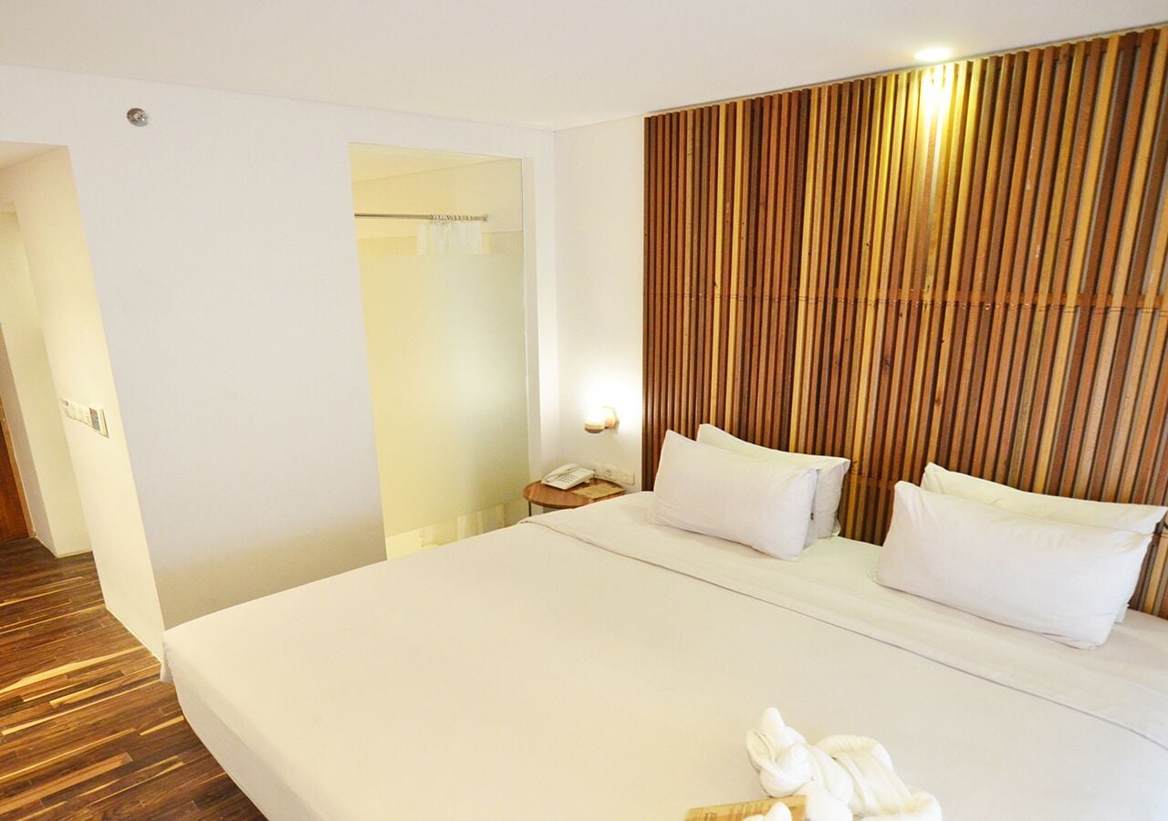 A hotel room with a large bed, a wooden headboard, a bedside table with a lamp, and a door leading to another room.