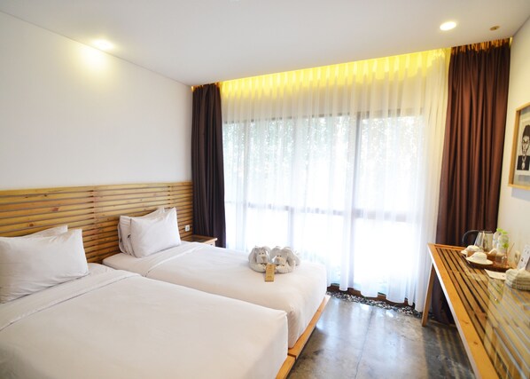 Habitación de hotel con dos camas, cabecera de madera, una mesita con una silla y un ventanal con cortinas.