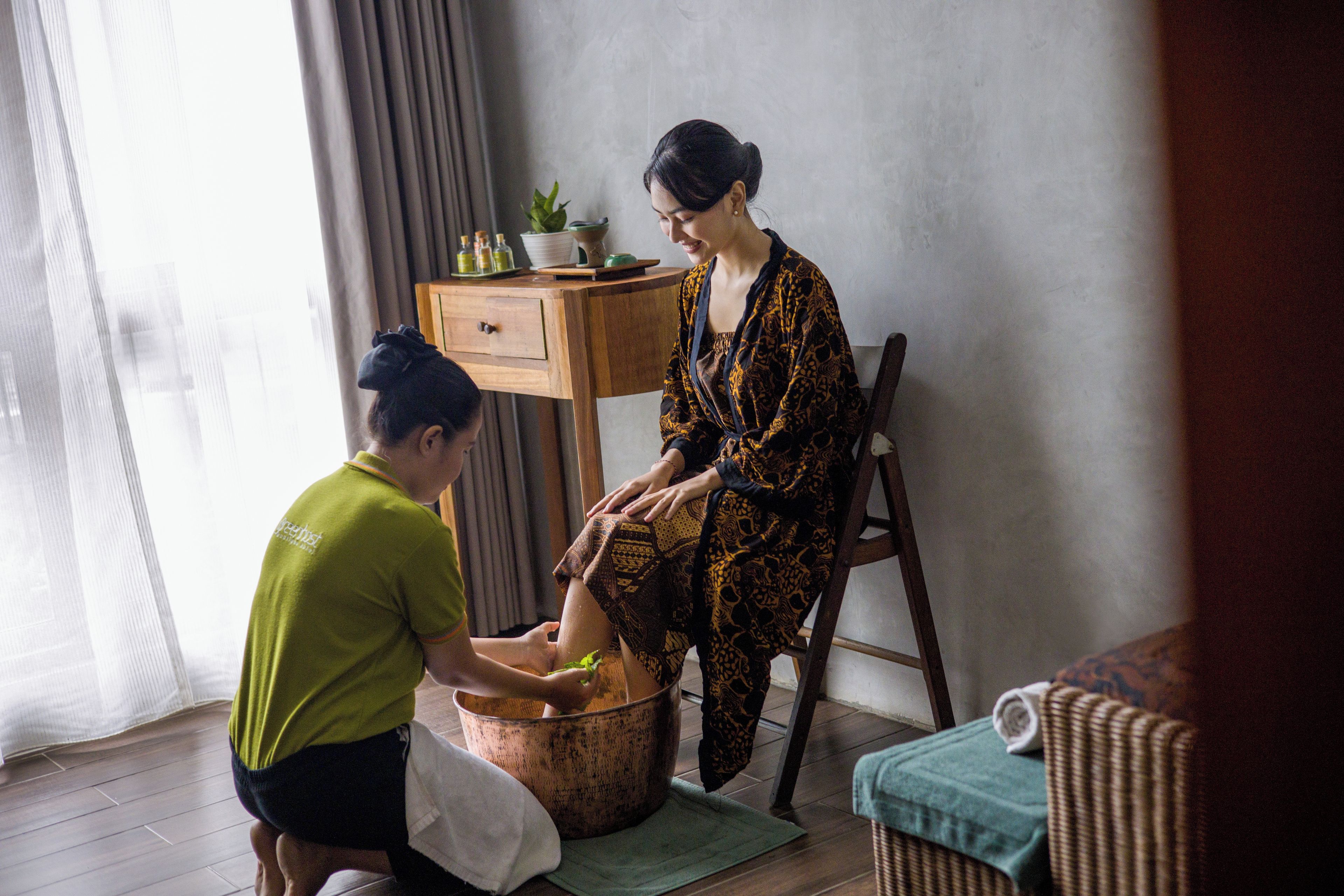 A person seated on a chair, another person kneeling beside them, and a wooden cabinet with a plant on top.