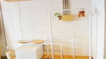 A minimalist room with a white brick wall, a wooden floor, a white radiator, a white chair, a small table with a plant, and a shelf with wooden utensils.