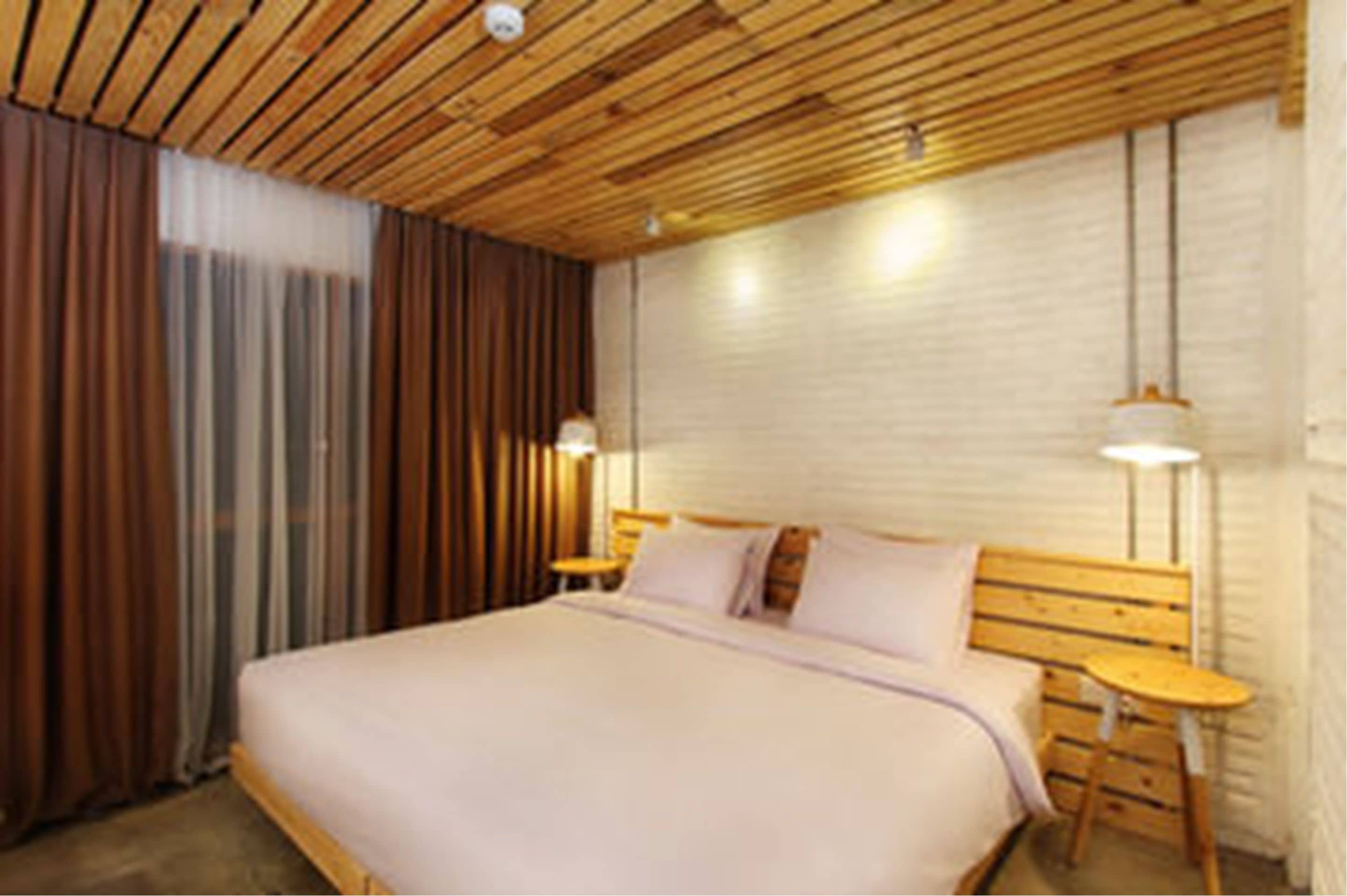 A bedroom with a wooden ceiling, a bed with a wooden headboard, two bedside tables, and a wall-mounted light fixture.