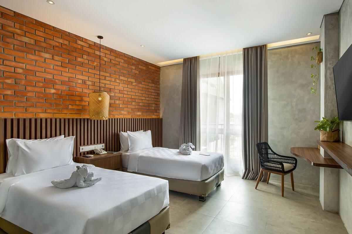 A hotel room with two beds, a desk, a chair, a TV, and a plant.