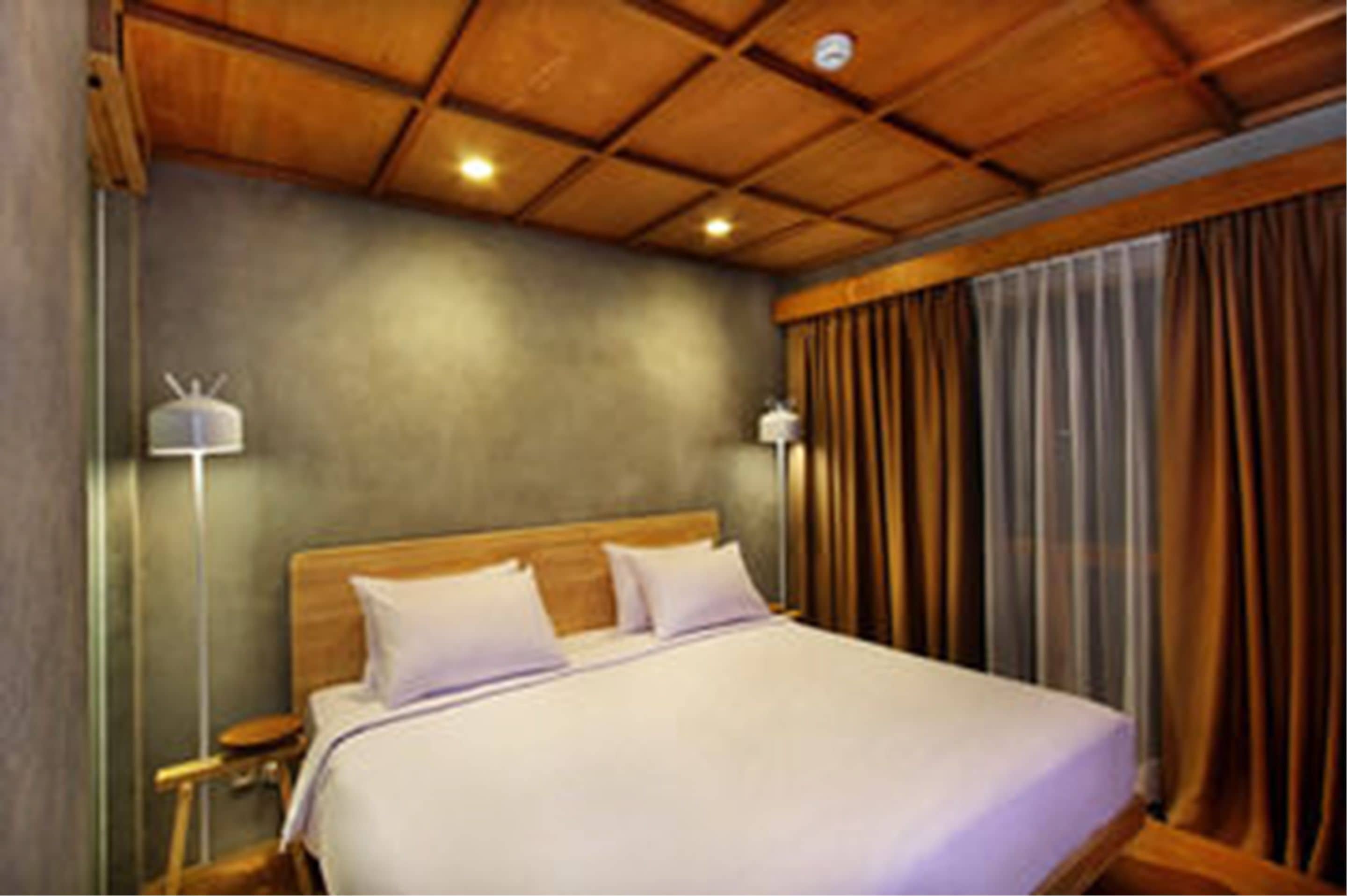 A hotel room with a wooden ceiling, a large bed with white linens, two bedside lamps, and a view of the outside through curtains.