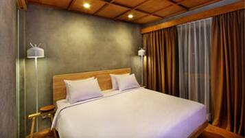 A hotel room with a wooden ceiling, a large bed with white linens, two bedside lamps, and a view of the outside through curtains.